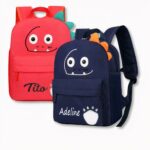 2 mochilas infantiles, una roja y otra azul oscuro con caras sonrientes de dinosaurios personalizadas con los nombres de Tito y Adeline, sobre fondo gris claro