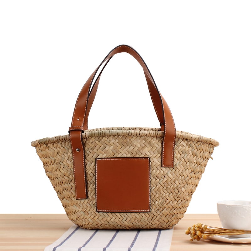 bolso de playa de paja con correa de cuero marrón y una pieza cuadrada de cuero en el lateral